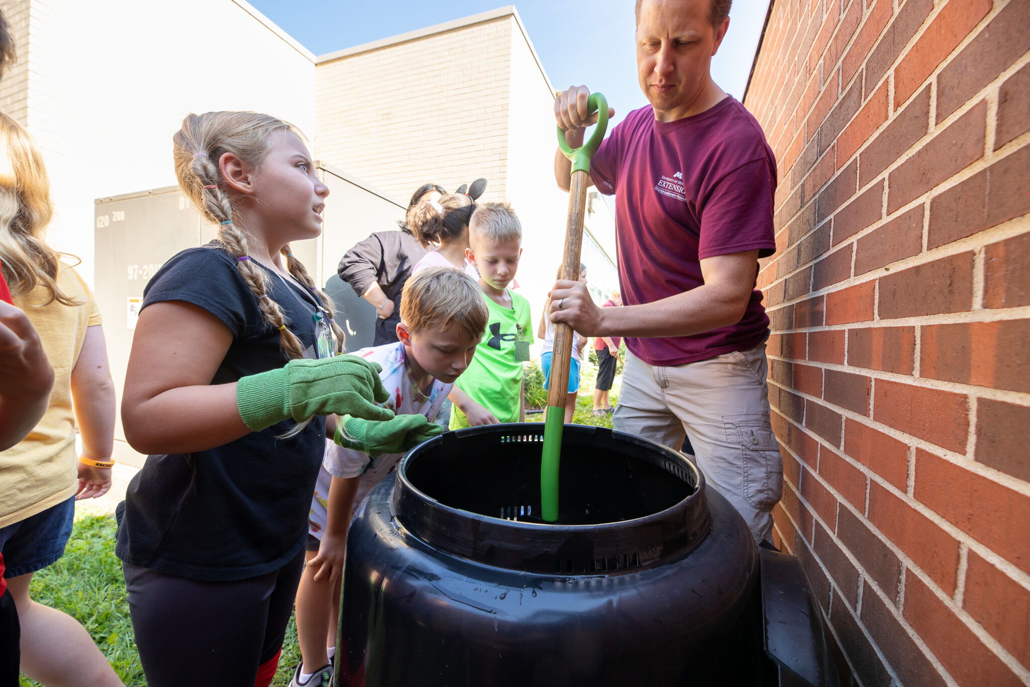 A man stirring a compost bin with children watching.