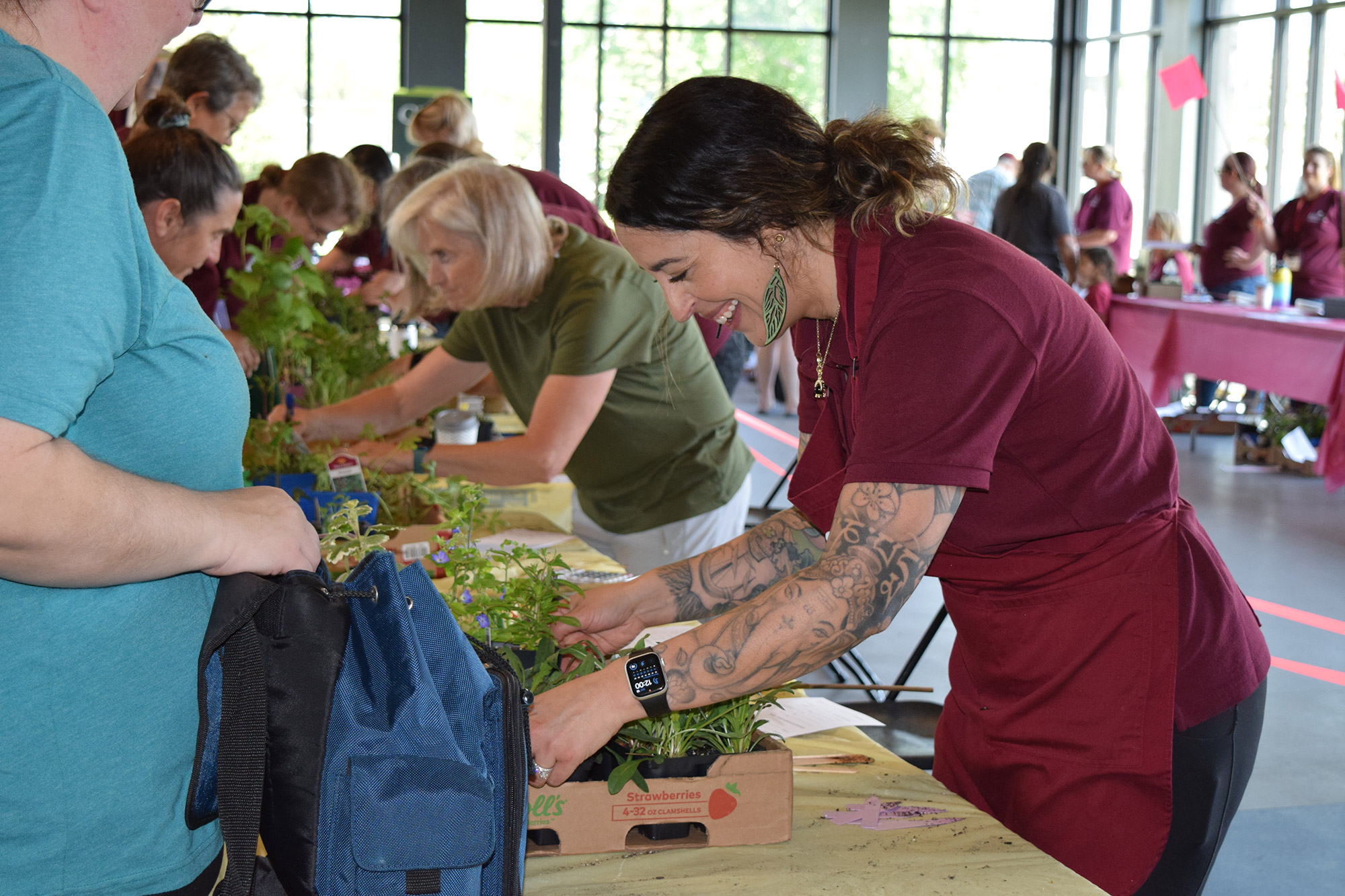 Volunteers arrange plants and flowers at plant sale, with focus on a woman in a maroon shirt and apron smiling and working with her hands.